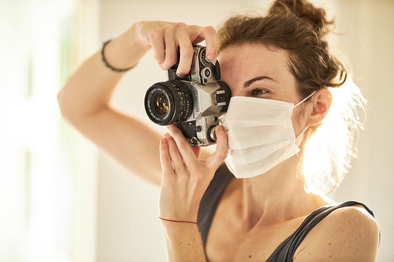 eine junge Frau mit Maske, die eine Spiegelreflexkamera im Anschlag hat.