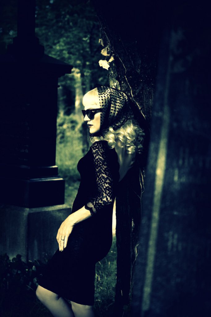 Frau mit Sonnenbrille und Spitzenkopftuch in kurzem schwarzen Kleid an einen Baum gelehnt. Im Hintergrund Grabsteine.