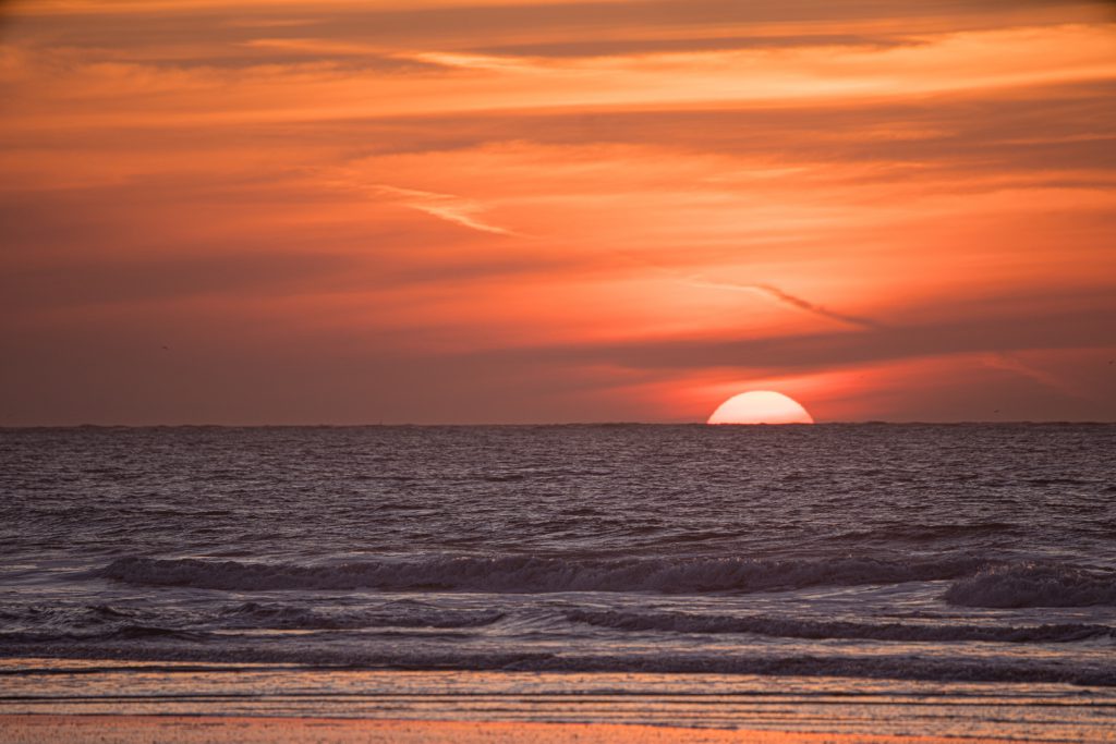 Im Vordergrund dunkelblaues wogendes Meer dahinter eine halbe weiße Sonnescheibe die ins Meer versinkt, im Hintergrund ein orangefarbener mit Streifen durchzogener wolkiger Himmel