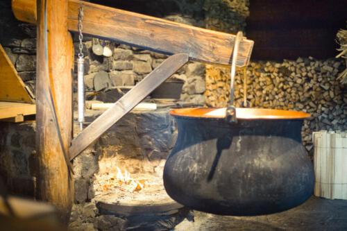 von Jürgen Wissel, im Vordergrund ist ein großer Kessel, der an einem schwenkbaren Holzbalken hängt, im Hintergrund eine Feuerstelle und an der Wand sind Holzscheide aufgestapelt.   