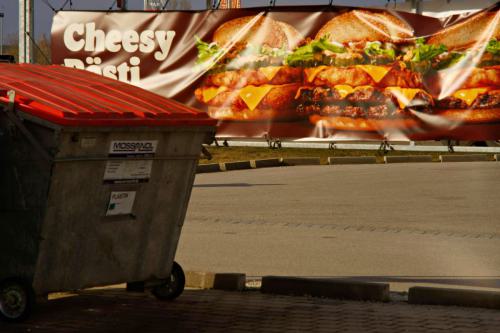 im Hintergrund eine Werbung eines Fastfoodresturant, im Vordergrund eine große Abfallbox.
